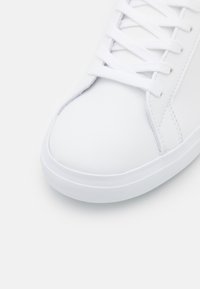 Sneaker bianca con tomaia in sintetico liscio, contraddistinta da lacci piatti, punta arrotondata e suola bianca testurizzata.