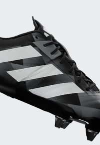 Svart och grå fotbollssko med svarta snören och tre vita diagonala ränder på sidan, med metallpiggar på sulan.