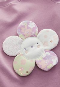 Un tissu violet présente une grande appliqué de fleur avec des pétales pastel aux motifs floraux variés, un visage souriant et des accents de sequins.