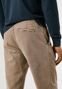Personne portant un pantalon beige et une chemise à manches longues foncée, vue de dos avec la main sur le côté et les détails des poches visibles.