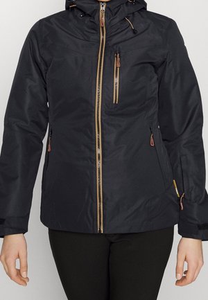 Mujer con chaqueta negra con capucha, detalles de cremalleras marrones y bolsillos con cremallera en el pecho y los lados, posando frente a un fondo liso.