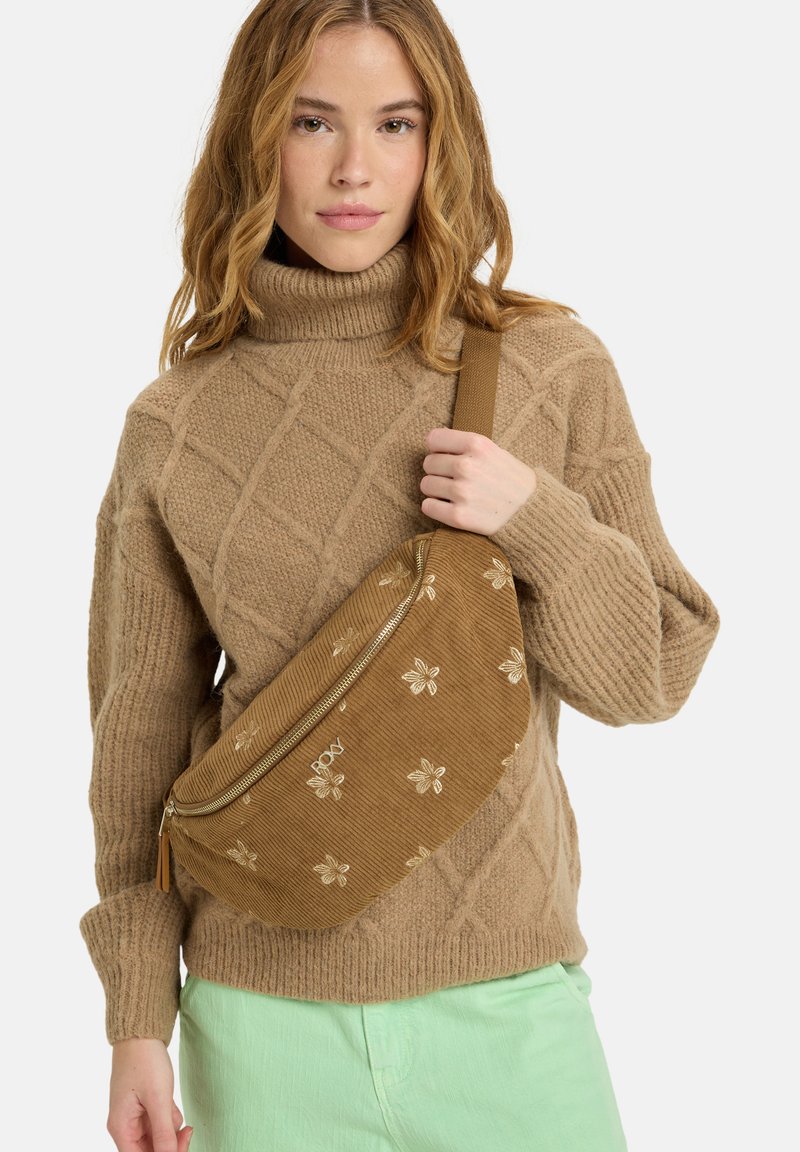 Braune Cordura-Slingtasche mit goldener Blumenstickerei, Reißverschluss und verstellbarem Tragegurt, getragen über einem beigen Kabelstrickpullover.