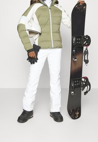 Snowboarder che indossa una giacca imbottita verde oliva e bianca, pantaloni bianchi, guanti neri e stivali neri, mentre tiene una tavola da snowboard.
