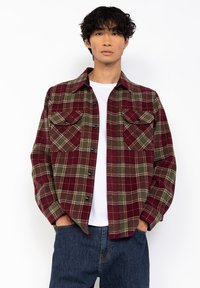 Czerwono-zielona koszula flanelowa w krateczkę z dwoma kieszeniami na piersi i czarnymi guzikami, noszona z białym T-shirtem i niebieskimi dżinsami.