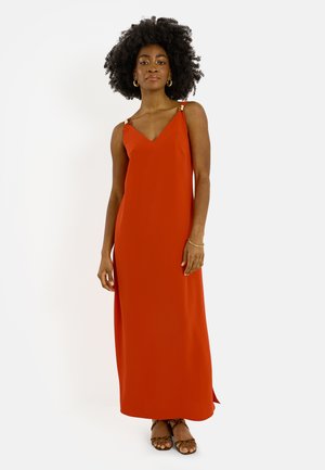 Mujer con cabello rizado natural, vestida con un vestido largo sin mangas de color naranja óxido y sandalias marrones, de pie frente a un fondo liso.