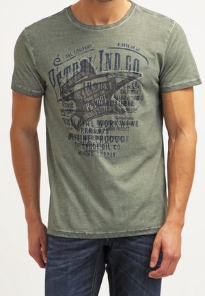 Mand iført falmet grøn kortærmet t-shirt med mørkeblå grafisk tekst og stjernedesign, kombineret med mørkeblå jeans.