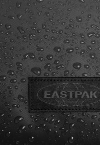 Tela impermeable negra con gotas de agua. Presenta una superficie texturizada y un parche rectangular del logo de Eastpak en la esquina inferior.