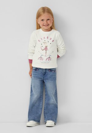 Weißer Sweatshirt mit "CIRCUS"-Druck und abgebildetem Tänzer; kombiniert mit hellblauen, weit geschnittenen Jeans mit bestickten Details. Weiße Sneaker.
