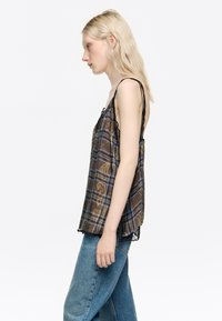 Brązowa kamizelka w kratę z czarną koronką, z cienkimi ramiączkami. Noszona z jeansami typu high-waist w niebieskim kolorze. Widok z profilu.