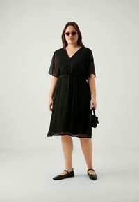Black, knee-length dress with short, sheer sleeves and button detail. Features an elastic waist and a flowy silhouette. Worn with flat shoes.
