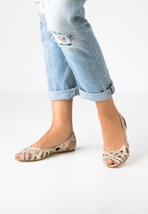 Beżowe płaskie buty z plecionym wzorem i odkrytymi palcami, z wycięciami. W połączeniu z podwiniętymi jasnoniebieskimi dżinsami.
