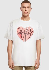 Hvid bomulds t-shirt med et stort hjertegrafik og teksten "SPREAD LOVE" i nuancer af pink og rød. Kortærmet og afslappet pasform.