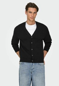 Cardigan nero con scollo a V, chiusura frontale con bottoni e maniche lunghe, indossato sopra una t-shirt bianca, abbinato a jeans azzurri.