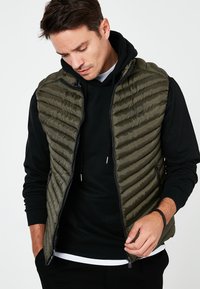 Olīvu zaļš pūkains veste ar horizontālu selēdējumus, ar rāvējslēdzēju un melnu kapuci kreklu zem tā. Tekstūru audums ar spīdīgu apdari.
