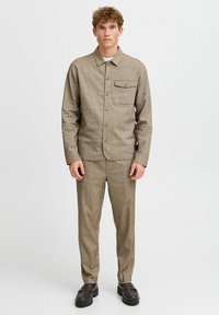 Young man wearing a matching brown button-up shirt jacket and pants, with black chunky loafers, standing against a plain white background.