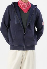 Persona che indossa una felpa con zip blu navy con un patch della bandiera americana, una bandana rossa intorno al collo, una maglietta nera e pantaloni beige, in piedi con le mani dietro la schiena.