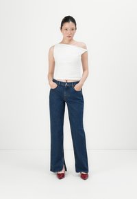 Kvinne med en asymmetrisk hvit topp, mørkeblå vide jeans med frontslisser, og røde spisse leppestifter, står mot en hvit bakgrunn.