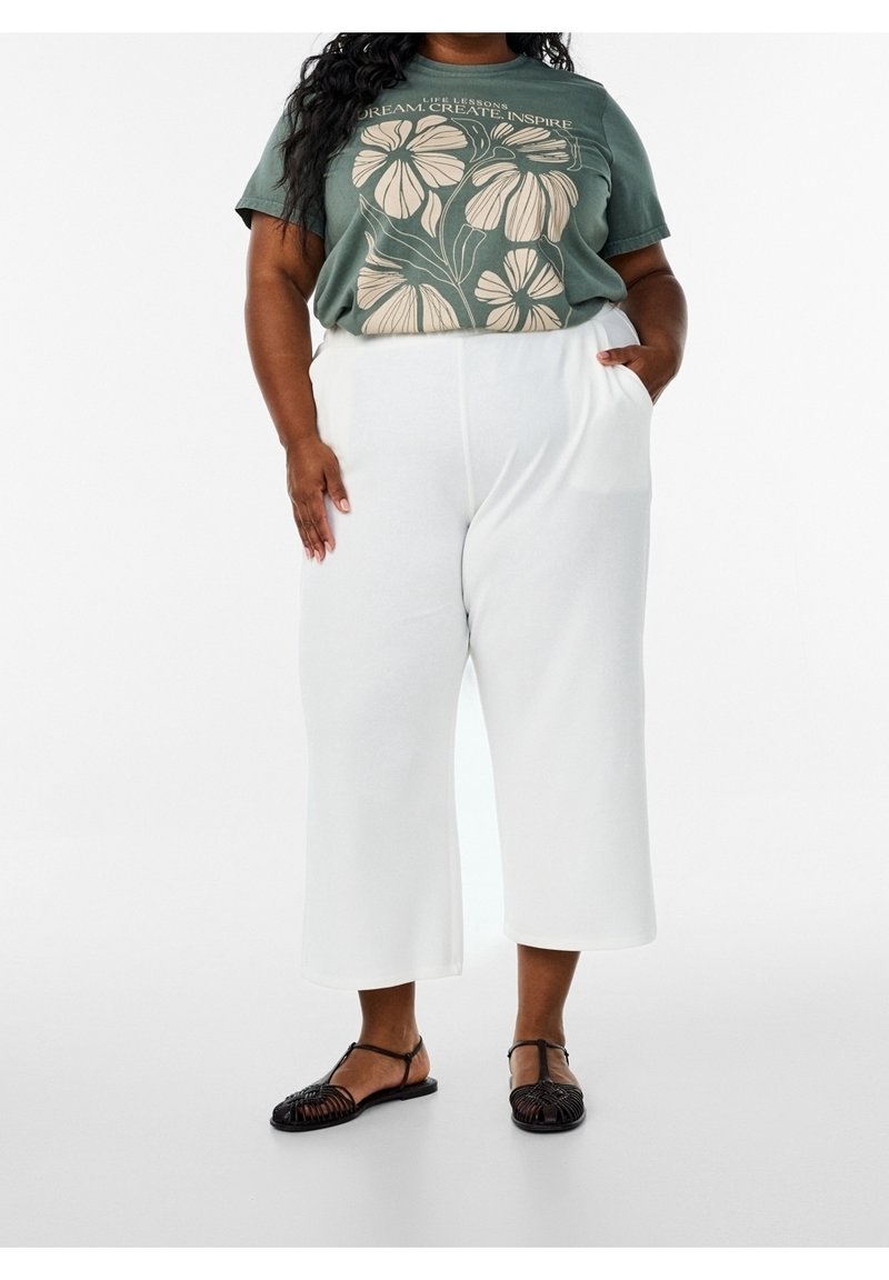 Femme portant un t-shirt graphique vert avec des fleurs et un pantalon blanc large court, debout avec une main dans la poche et des sandales noires.