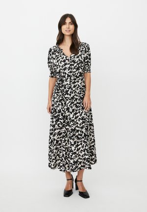 Schwarz-weißes Kleid mit Blattmuster, V-Ausschnitt, kurzen Puffärmeln und fließender Maxi-Länge, kombiniert mit schwarzen Schuhen mit Blockabsatz.