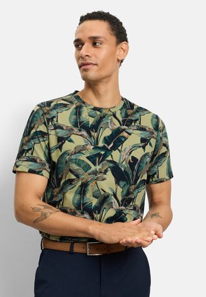 Mann mit kurzen lockigen Haaren, der ein grünes T-Shirt mit tropischem Blattmuster, marineblaue Hose, braunen Gürtel und Vogel-Tattoos auf beiden Unterarmen trägt.