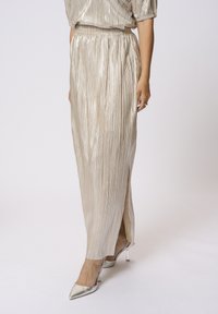Jupe midi plissée métallisée couleur champagne avec fente latérale, taille élastique et texture douce, associée à des talons argentés brillants.