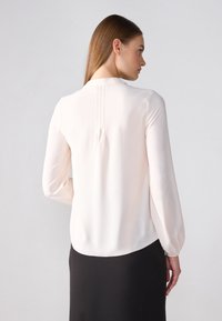 Blusa de manga larga rosa claro, textura de satén suave, dobladillo redondeado, detalle de pliegue en la espalda, puños con botones, combinada con una falda negra.