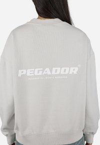 Light gray sweatshirt made of cotton, featuring a ribbed neckline and cuffs. Large white "PEGADOR" logo printed on the back.