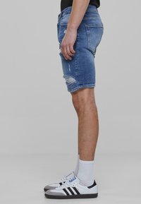 Denim šorti, gaiši zili, vidējas garuma, ar saplēstām malām un nodilušiem detalēm. Valkāti ar baltiem un melniem kedām un baltām zeķēm.