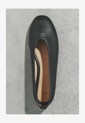 Black leather flat shoe with rounded toe and tan insole placed on light gray textured surface.