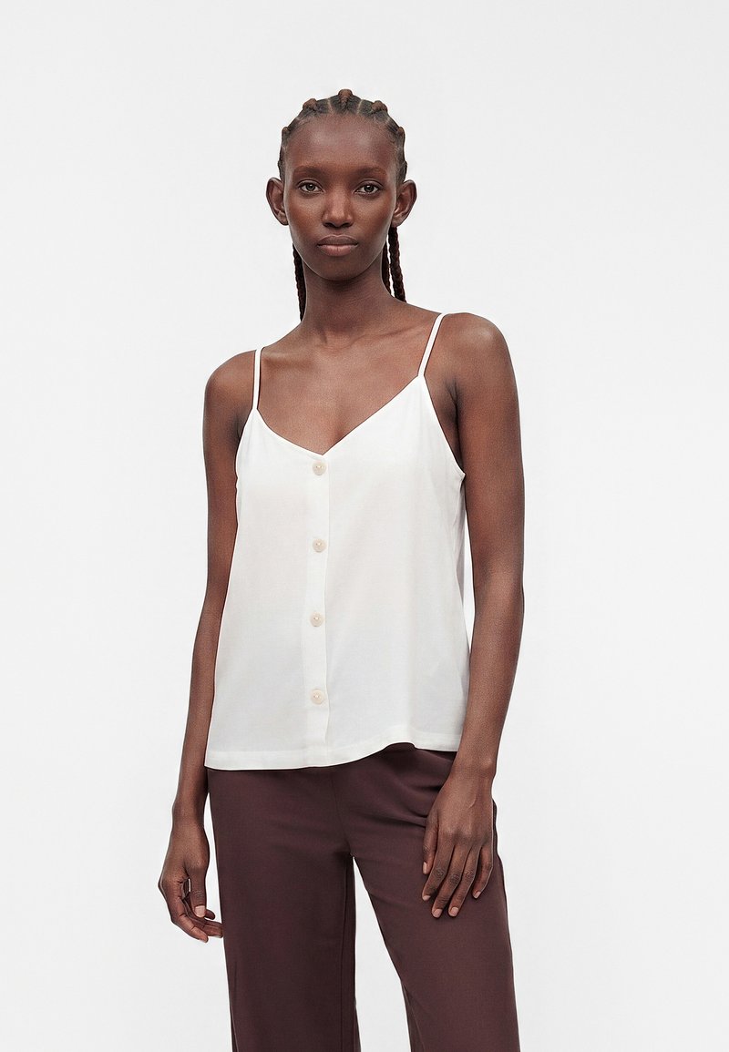 Femme aux cheveux tressés portant un haut blanc sans manches boutonné et un pantalon marron, debout devant un fond clair uni.