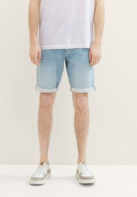 Pantalones cortos de mezclilla azul claro con dobladillos enrollados, con un corte clásico. Combinados con zapatillas blancas y beige, con un fondo neutro.