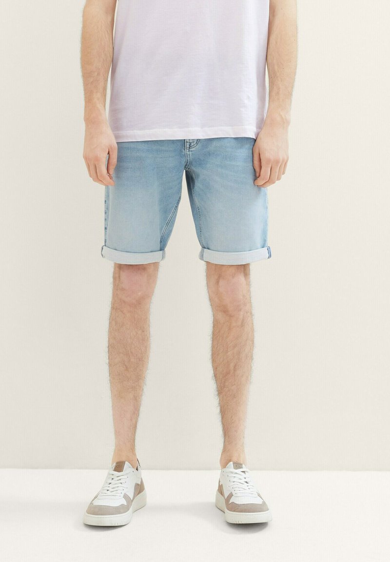 Pantalones cortos de mezclilla azul claro con dobladillos enrollados, con un corte clásico. Combinados con zapatillas blancas y beige, con un fondo neutro.