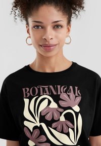 Zwarte katoenen t-shirt met een botanische afbeelding in crème en gedempt roze. Korte mouwen, ronde halslijn, zachte textuur.