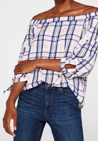 Schulterfreies Karomuster-Oberteil in Weiß, Marineblau und Rosa. Mit aufgerollten Ärmeln und lockerer Passform. Kombiniert mit dunkelblauen Denim-Jeans.