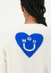 Pull en tricot blanc avec un grand motif de cœur bleu dans le dos, accompagné du texte blanc "HGO U" à l'intérieur du cœur. Tissu texturé.