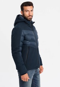 Navyblaue Jacke mit Kapuze, die eine gepolsterte Brust und einen glatten unteren Teil bietet. Verfügt über einen Reißverschluss und seitliche Taschen.
