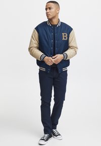 Jūrinės ir smėlio spalvos varsity striukė su snap mygtukais, įrengtais apykaklėmis ir laiško "B" emblema; derinama su tamsiomis kelnėmis ir juodais sportbačiais.