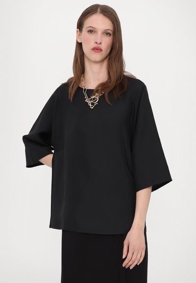Zwarte korte mouwen top met een losse pasvorm, ronde halslijn en zachte textuur. Gedragen met een decoratieve metalen ketting. Minimalistisch ontwerp.