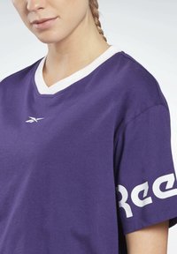 Fialové bavlněné tričko s výstřihem do V a bílým límečkem. Na levé straně hrudníku je bílá značka a na rukávu kontrastní bílý nápis.