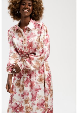 Uśmiechnięta kobieta z kręconymi włosami, ubrana w białą sukienkę z długim rękawem z różowo-brązowym kwiatowym wzorem i zapinanymi na guziki mankietami.