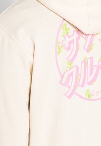 Gräddfärgad hoodie i bomull med en stor rund tryck i rosa och grönt, med japansk text och grafiska element.