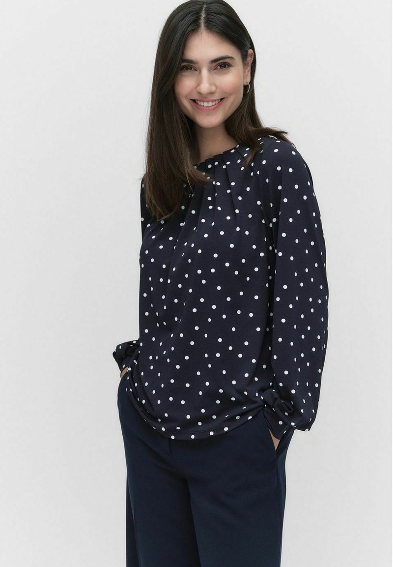 Navy blouse met witte stippen, zachte stof, ontspannen pasvorm, lange mouwen, geplooid detail bij de halslijn en zijzakken. Gecombineerd met donkere broek.