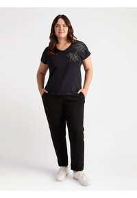 Czarna koszulka z dekoltem w kształcie V z ozdobnym srebrnym haftem kwiatowym na lewym ramieniu, zestawiona z czarnymi spodniami i białymi sneakersami.