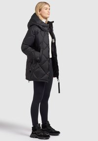 Doudoune matelassée noire avec capuche, dotée d'une fermeture éclair, de poches latérales et d'une étiquette de marque. Portée par-dessus un haut blanc et un legging noir.