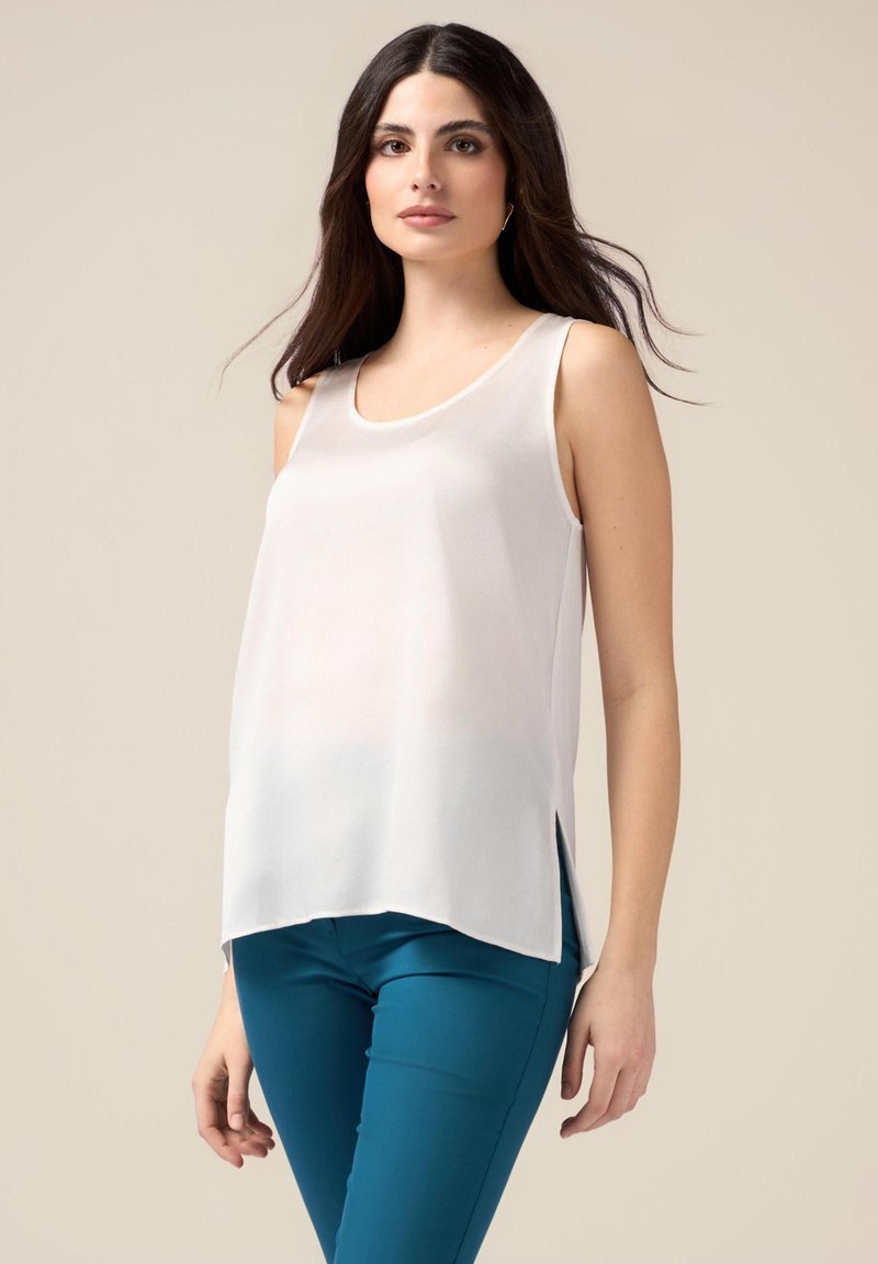 Top bianco senza maniche con scollo rotondo, caratterizzato da fessure laterali e una texture liscia e leggera, abbinato a pantaloni aderenti teal.