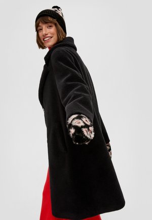 Manteau en polaire noir avec un col oversized et des poignets en peluche à motifs, porté sur un vêtement rouge. Présente une coupe décontractée et une fermeture à boutons.