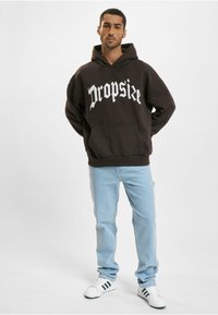 Brun hættetrøje med hvid gotisk "Dropsize" tekst, frontlomme, parret med lyseblå jeans og hvide sneakers med grønne striber.