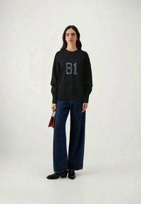 Svart sweatshirt med glittriga bokstäver "G U D E R 81" på framsidan, tillsammans med högmidjade, vida mörkblå jeans och svarta skor.