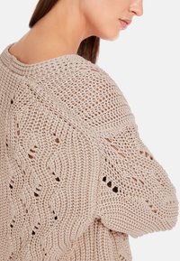 Hellbeiger, gestrickter Pullover mit einem lockeren, offenen Muster und breiten, strukturierten Ärmeln mit Seitenschlitzen. Glattes Garn mit einer weichen Optik.
