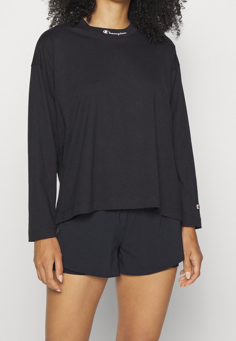 Femme portant un haut Champion noir à manches longues avec le logo sur le col et un short noir assorti, debout devant un fond clair uni.